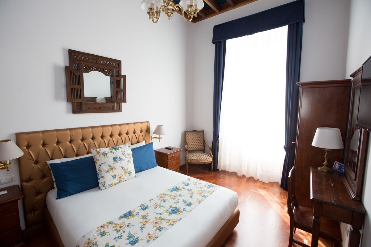 A hotel room with a large bed, a wooden nightstand, a chair, a desk, and a mirror.