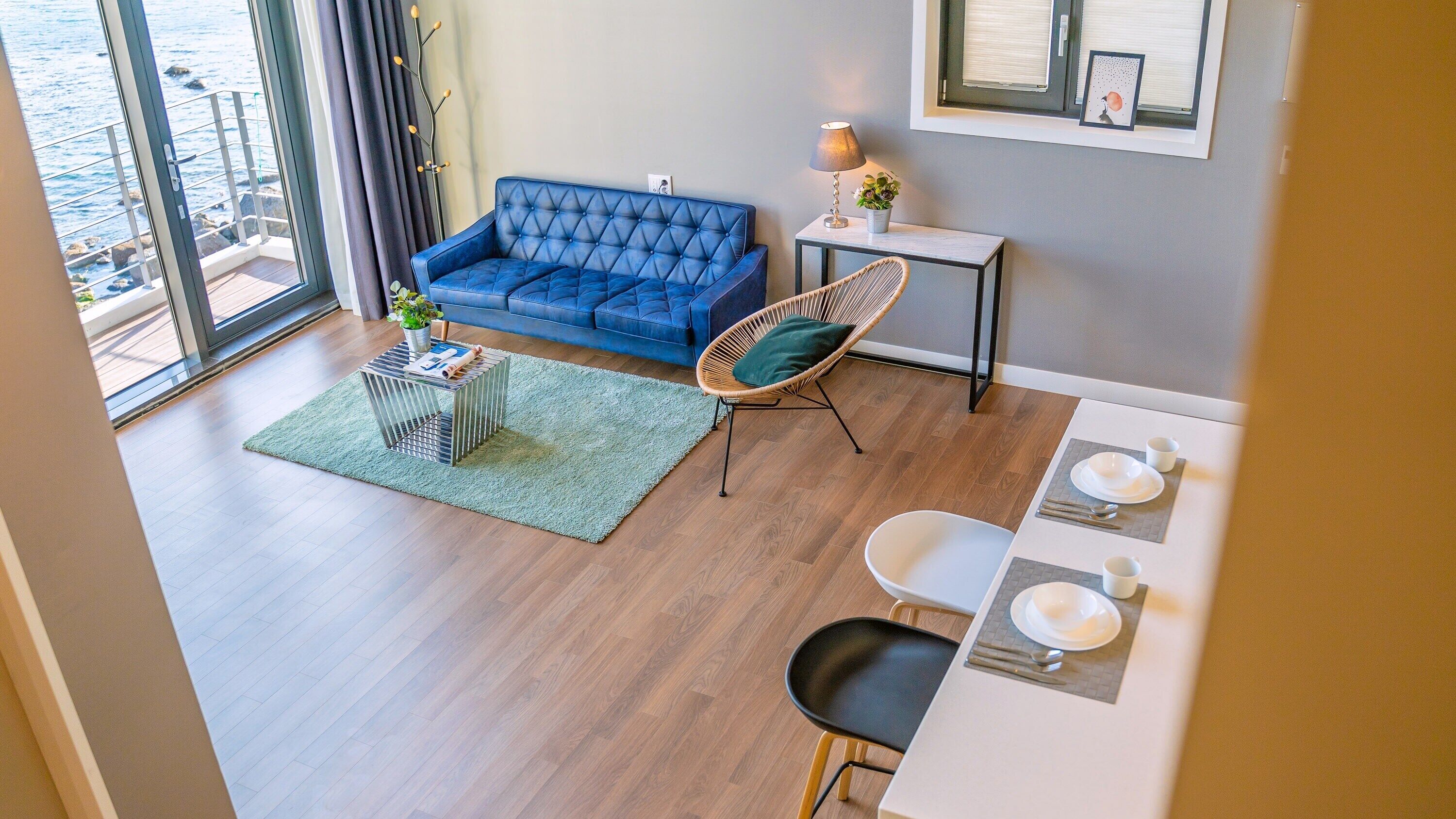 A modern living room with a blue sofa, a wicker chair, a dining table with chairs, and a view of the sea through large windows.