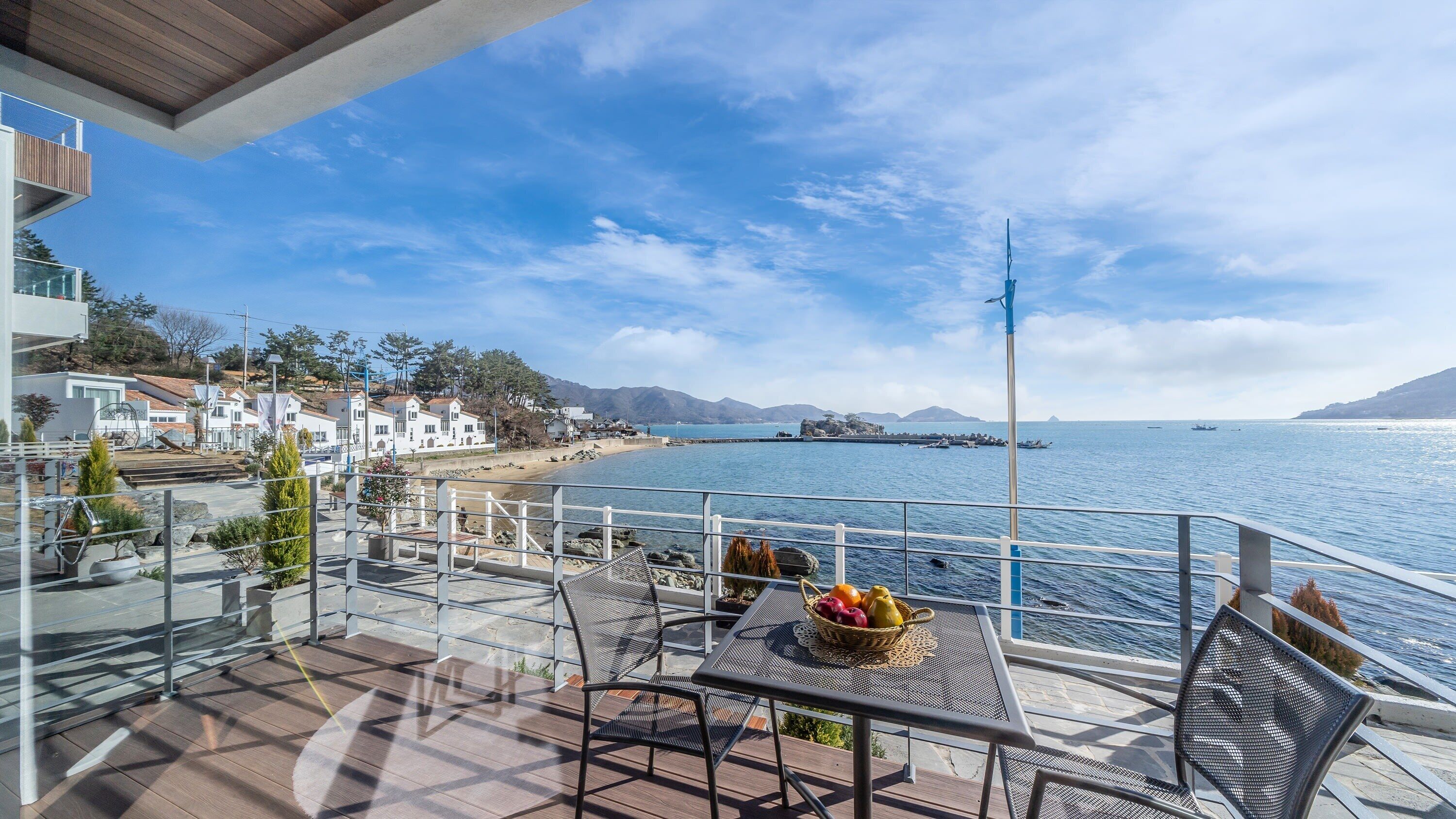 A balcony with a table and chairs overlooking a coastal area with buildings and a clear sky.