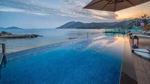 En infinitypool med utsikt över havet och bergen, ett trädäck och en loungehörna med en skylt som visar att den är öppen.
