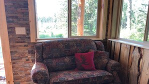 A cozy room with a patterned armchair, a wooden wall, and a window overlooking a forest. - CAM'S LAKESIDE BUNKHOUSE  (Soldotna)