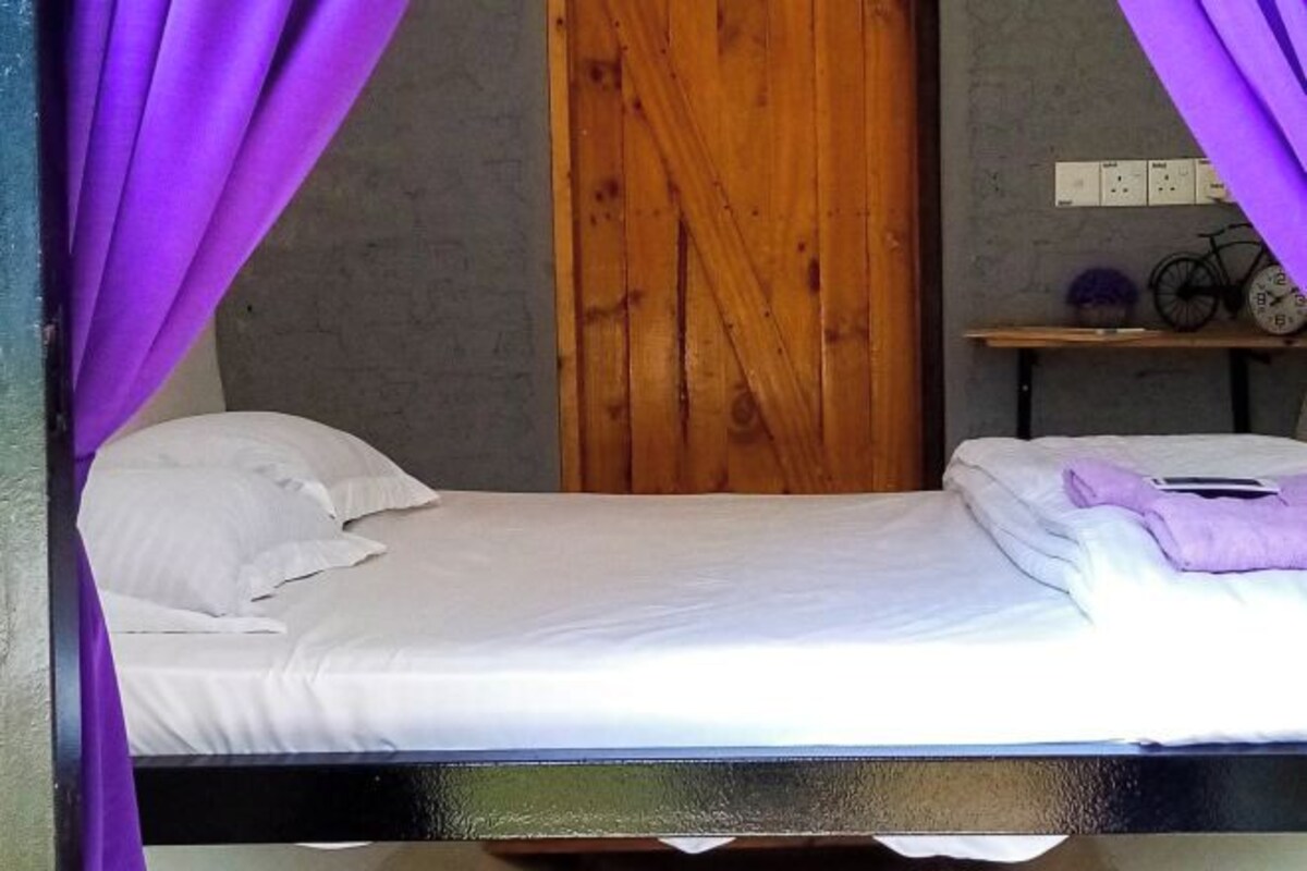 Una cama individual con ropa de cama blanca, una puerta de madera y una estantería con una bicicleta y un reloj.