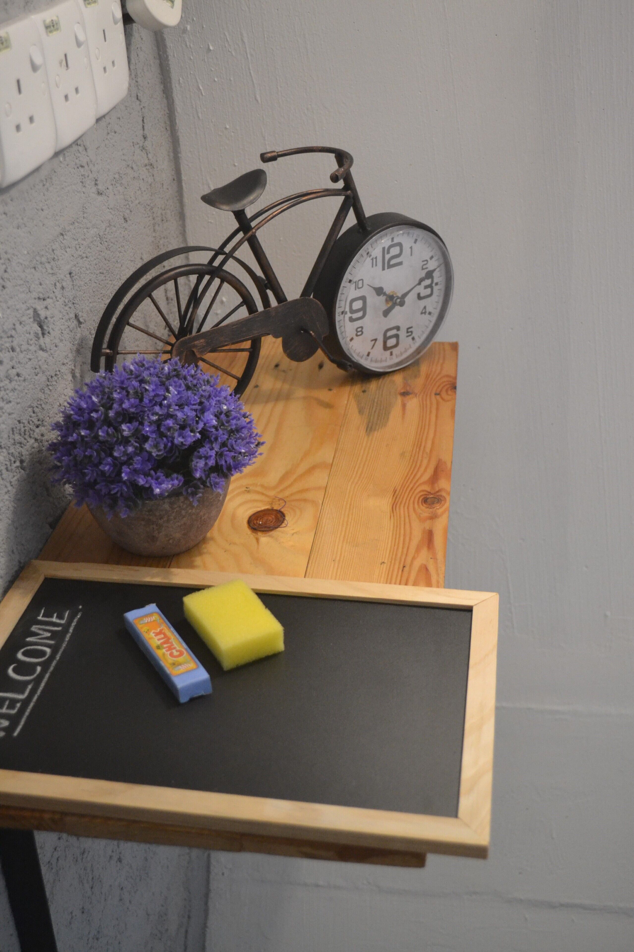 A wooden table with a chalkboard sign reading "WELCOME," a potted purple flower, a yellow sponge, and a black bicycle clock.