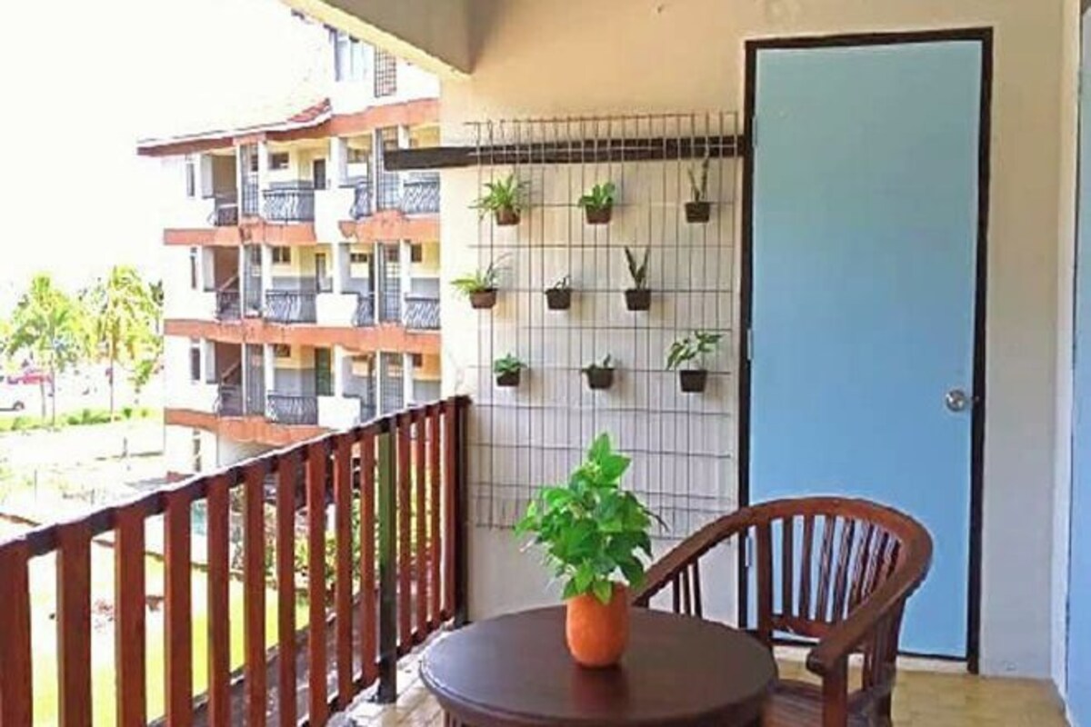 Un balcón con barandilla de madera, una mesa redonda de madera con una planta en maceta, y una puerta azul.
