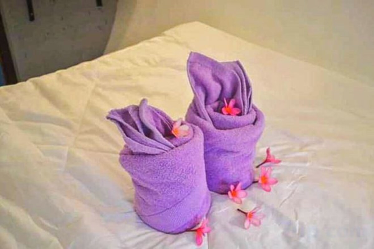 A bed with a white sheet and two purple towels folded into a shape resembling a gift, with a single pink flower placed on the bed.