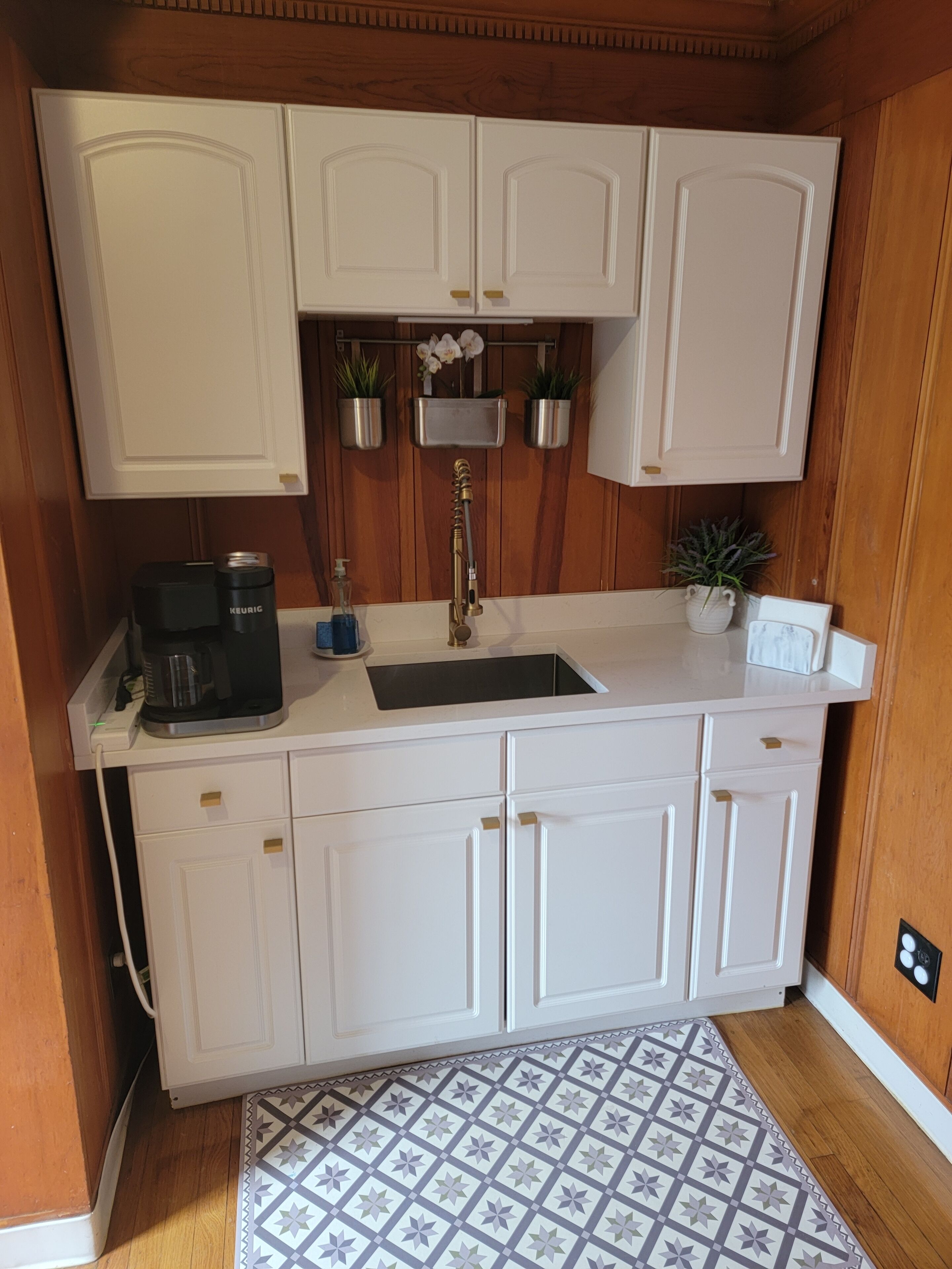 A compact kitchen with white cabinets, a built-in sink, a coffee maker, and potted plants.