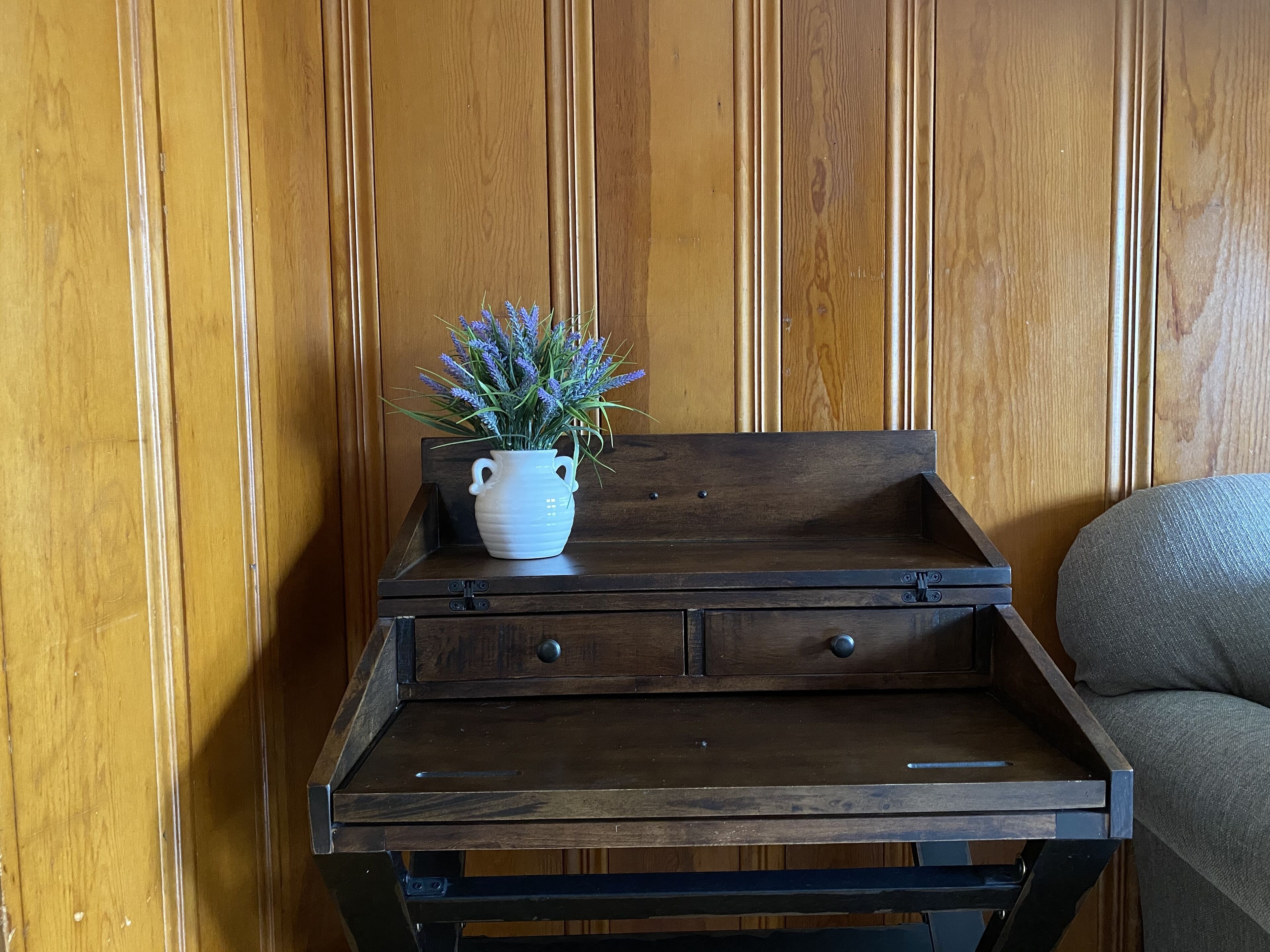 A wooden side table with a white vase containing lavender flowers, placed against a wooden paneled wall.