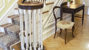 Eine imposante Treppe mit hölzernem Geländer und weißen Balustern, mit Teppich belegte Stufen und ein Sessel mit einem blumigen Kissen.
