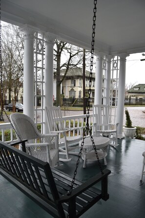 A porch with white wicker furniture, including a swing and chairs, under a white canopy with columns.
