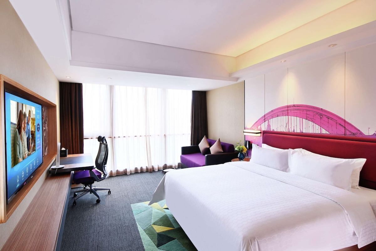 Quarto de hotel com uma cama grande, uma escrivaninha com cadeira, televisão na parede e janela com cortinas.