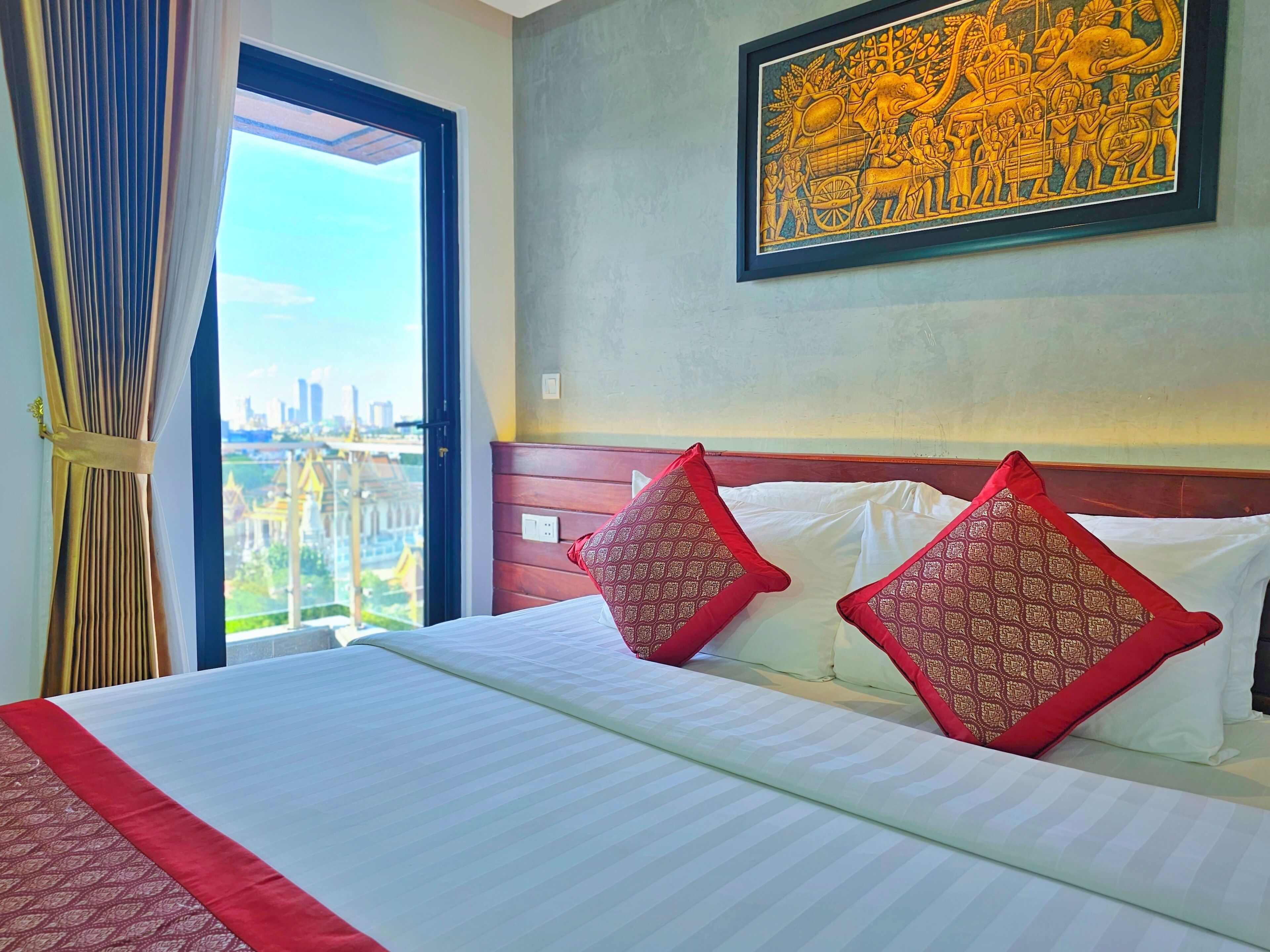 A hotel room with a large bed, two red patterned pillows, a view of the city through a window, and a framed artwork on the wall.