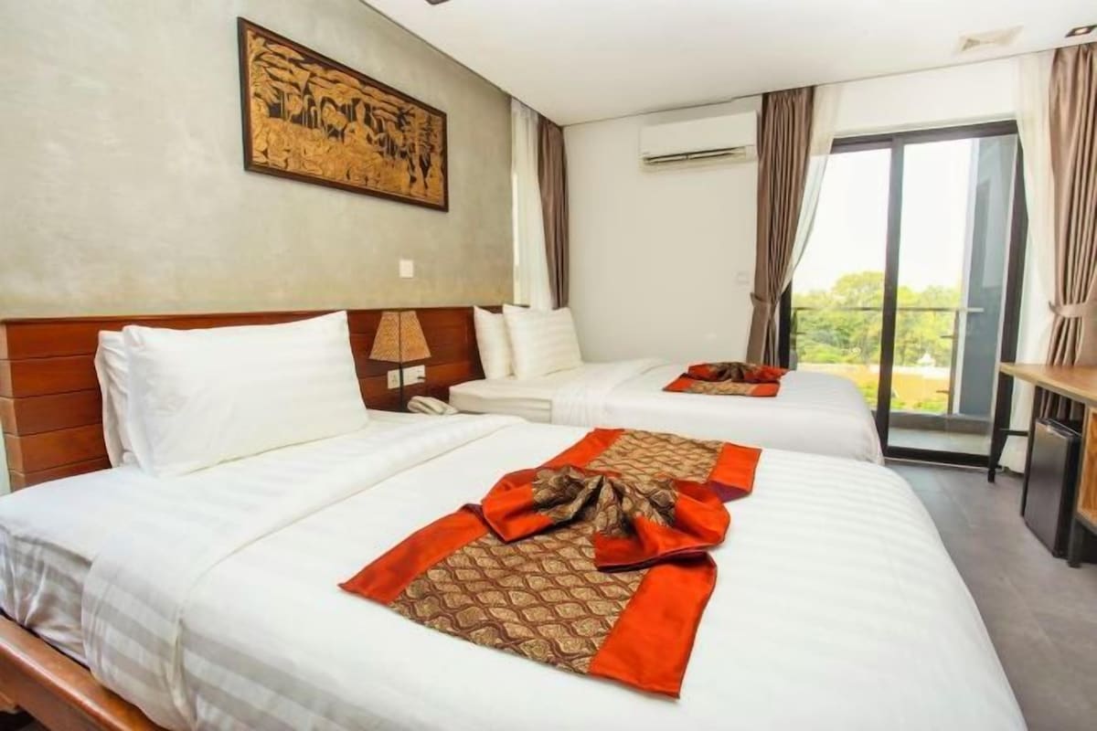 A hotel room with two beds, a wooden headboard, a painting on the wall, an air conditioning unit, and a view through the window.