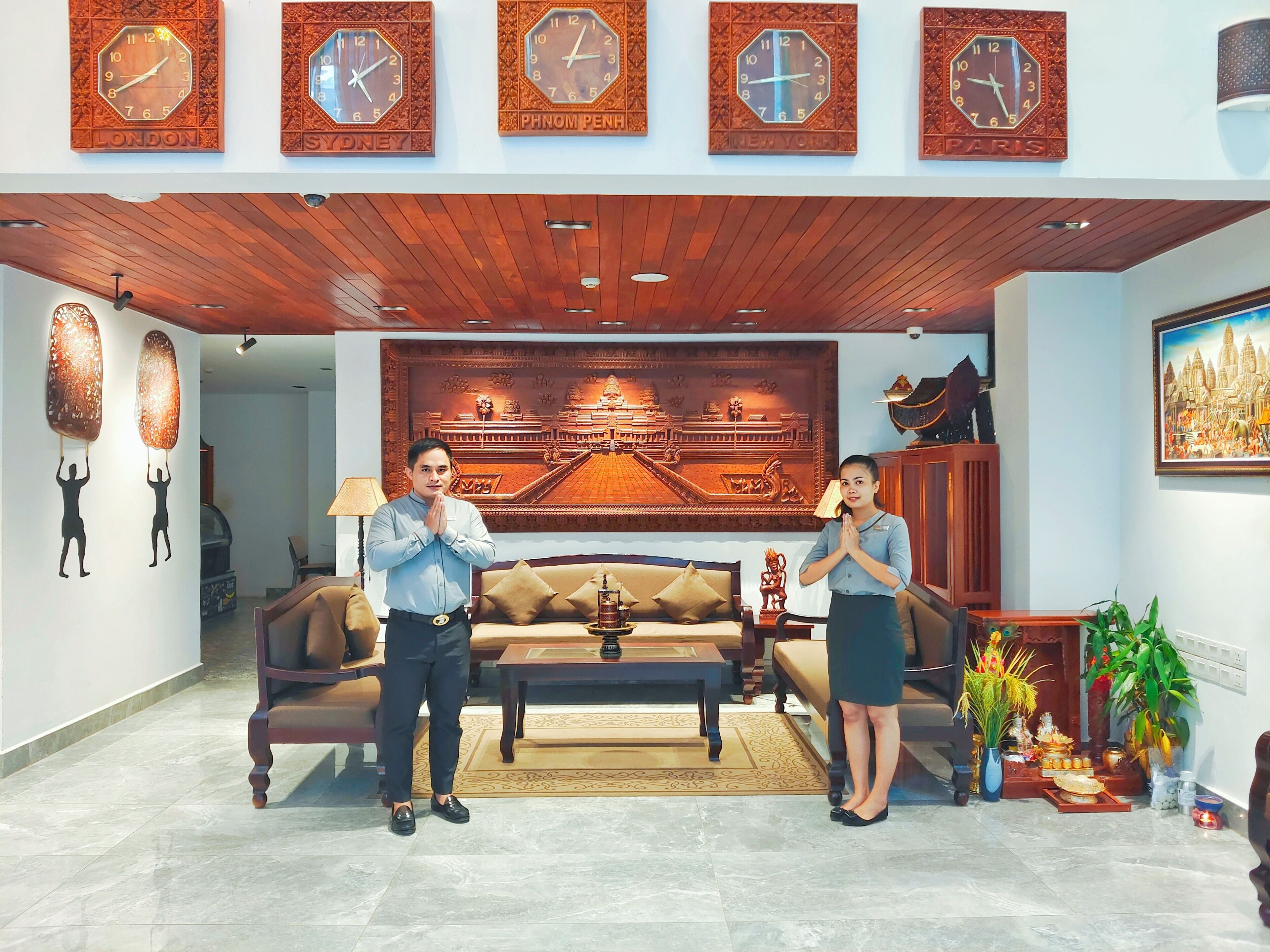 A hotel lobby with a wooden ceiling, a large Buddha statue, a sofa, and a decorative wall with clocks labeled 'LONDON', 'SYDNEY', 'PHNOM PENH', 'NEW YORK', and 'PARIS'.