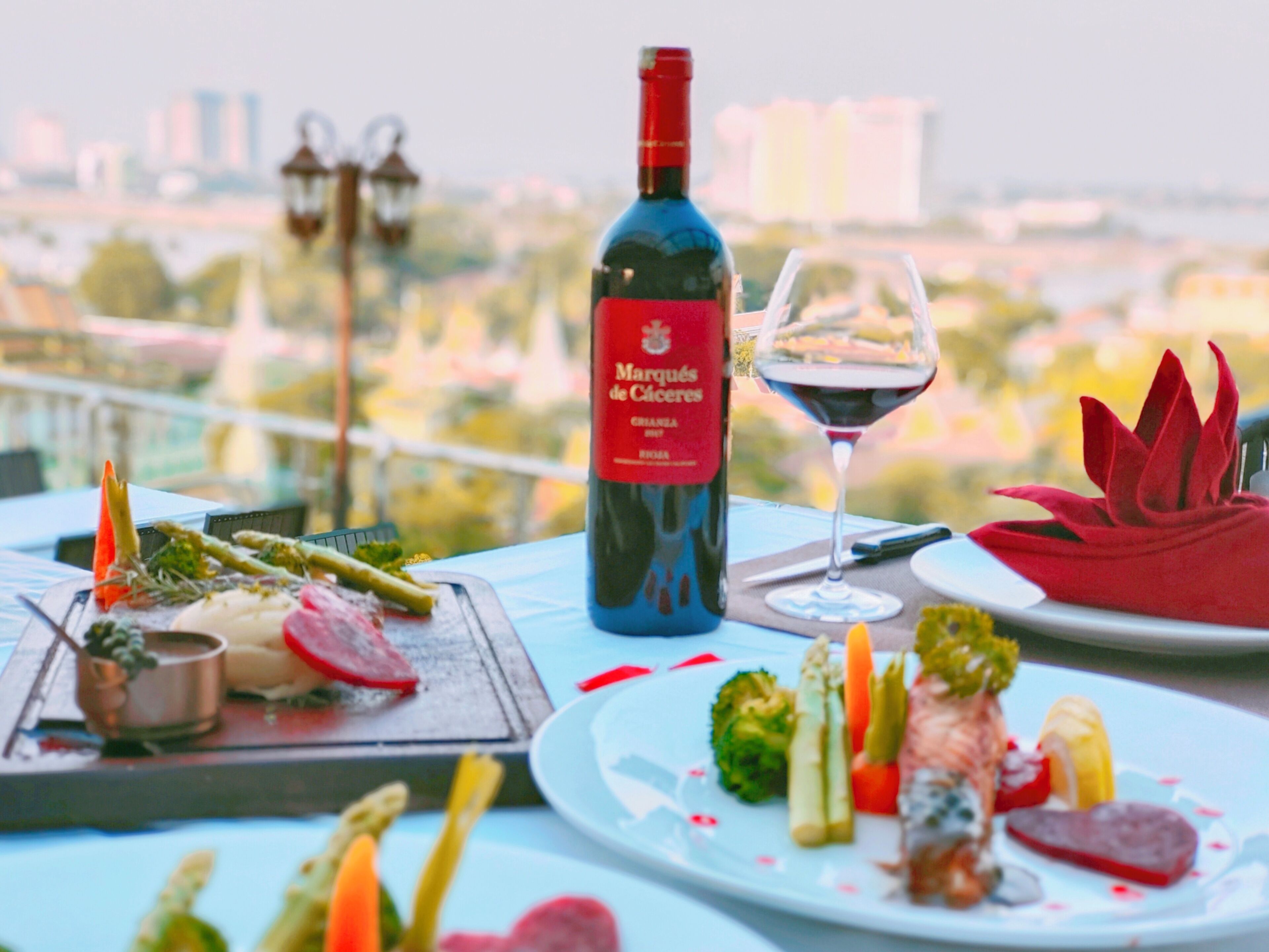 A table set with a bottle of wine, a glass of wine, and various dishes including asparagus, broccoli, and a red flower centerpiece.