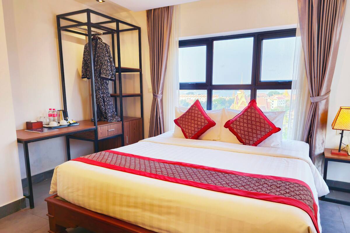 Quarto de hotel com uma cama grande, uma secretária de madeira, um guarda-roupa e uma janela com cortinas.