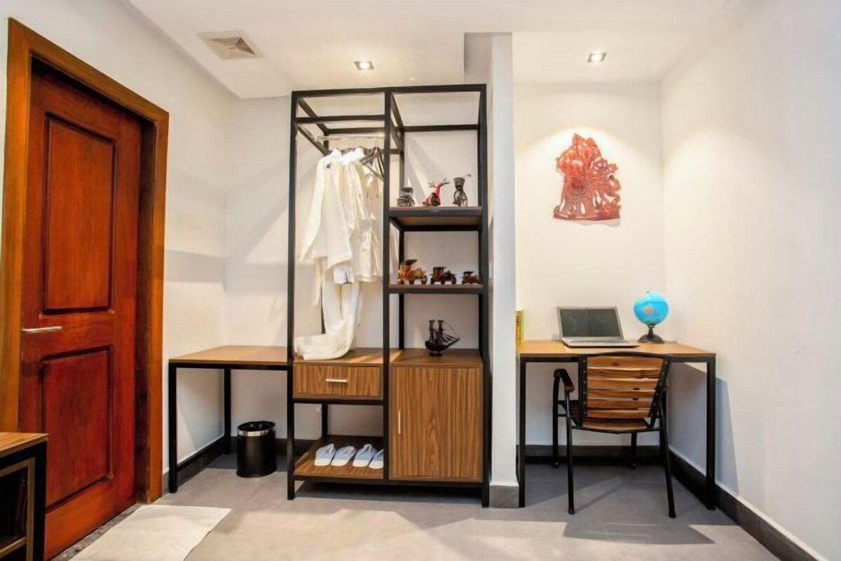 Une pièce de taille modeste, avec un bureau en bois, une chaise, un lit doté d’une structure en métal noir, une petite étagère contenant divers objets et une sculpture rouge accrochée au mur.