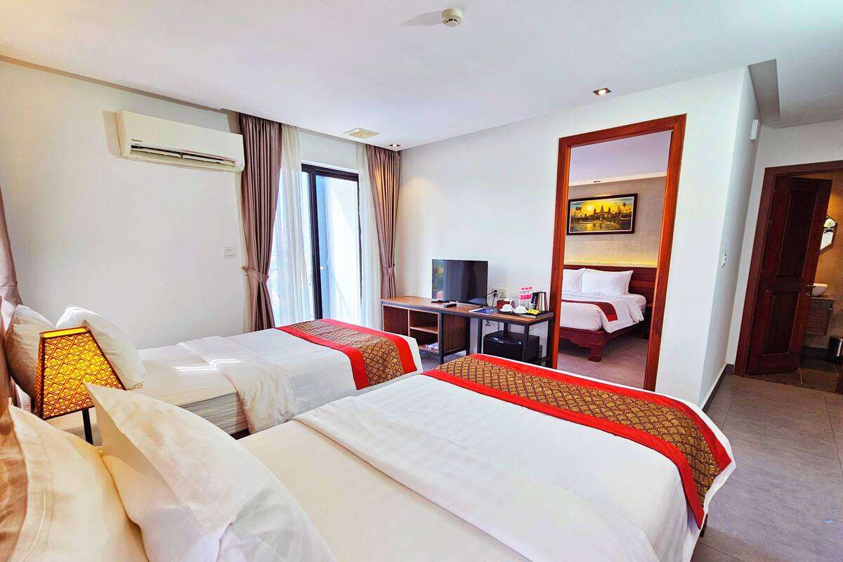 Quarto de hotel com duas camas, uma mesa de trabalho com televisão, um espelho e um banheiro visível por uma porta aberta.
