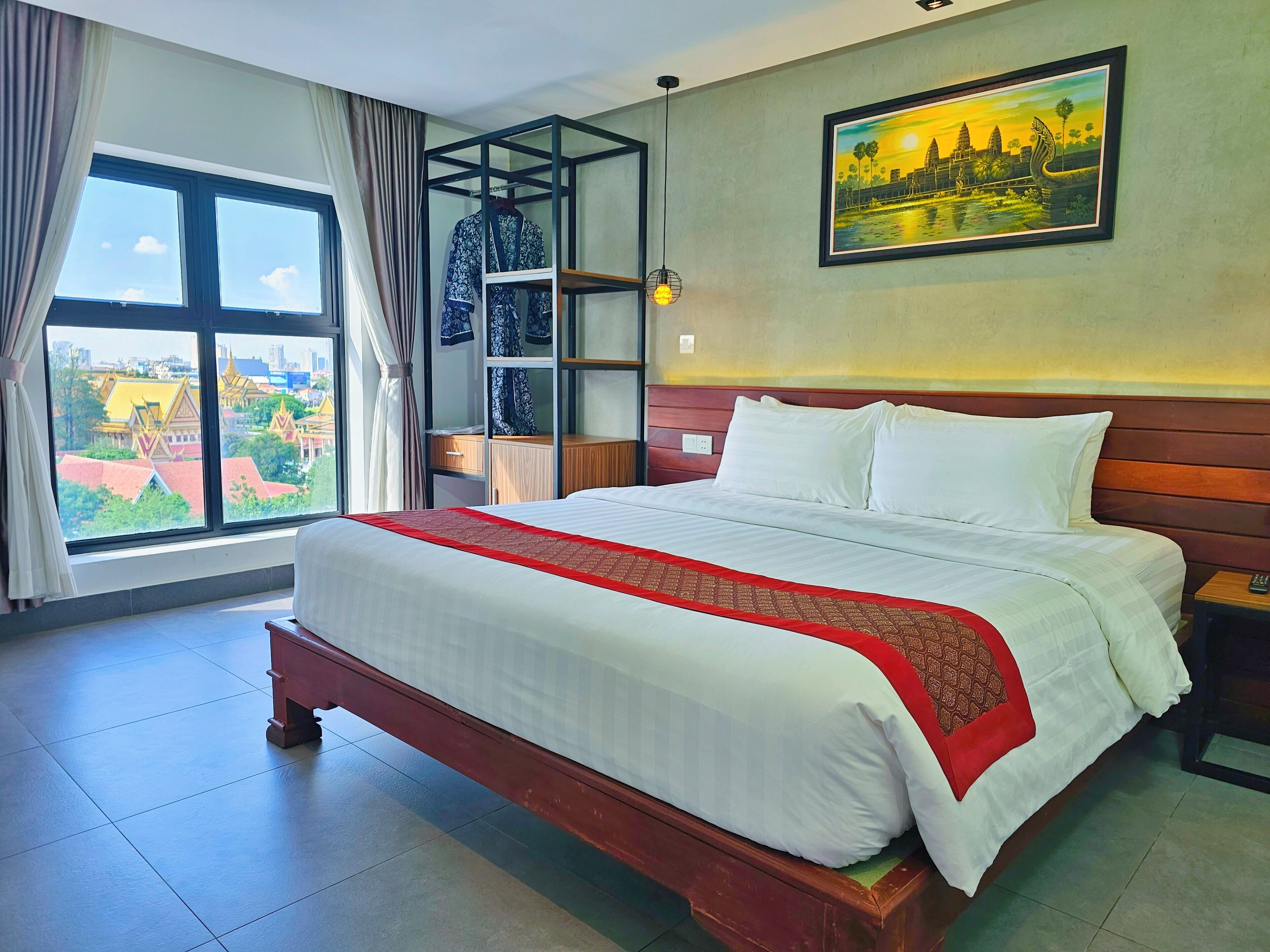 A hotel room with a large bed, a wardrobe, a nightstand, a painting, and a window with a view of a cityscape.