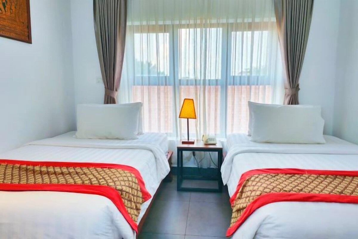 Une chambre d’hôtel avec deux lits simples, une table de chevet avec une lampe et des rideaux légers.