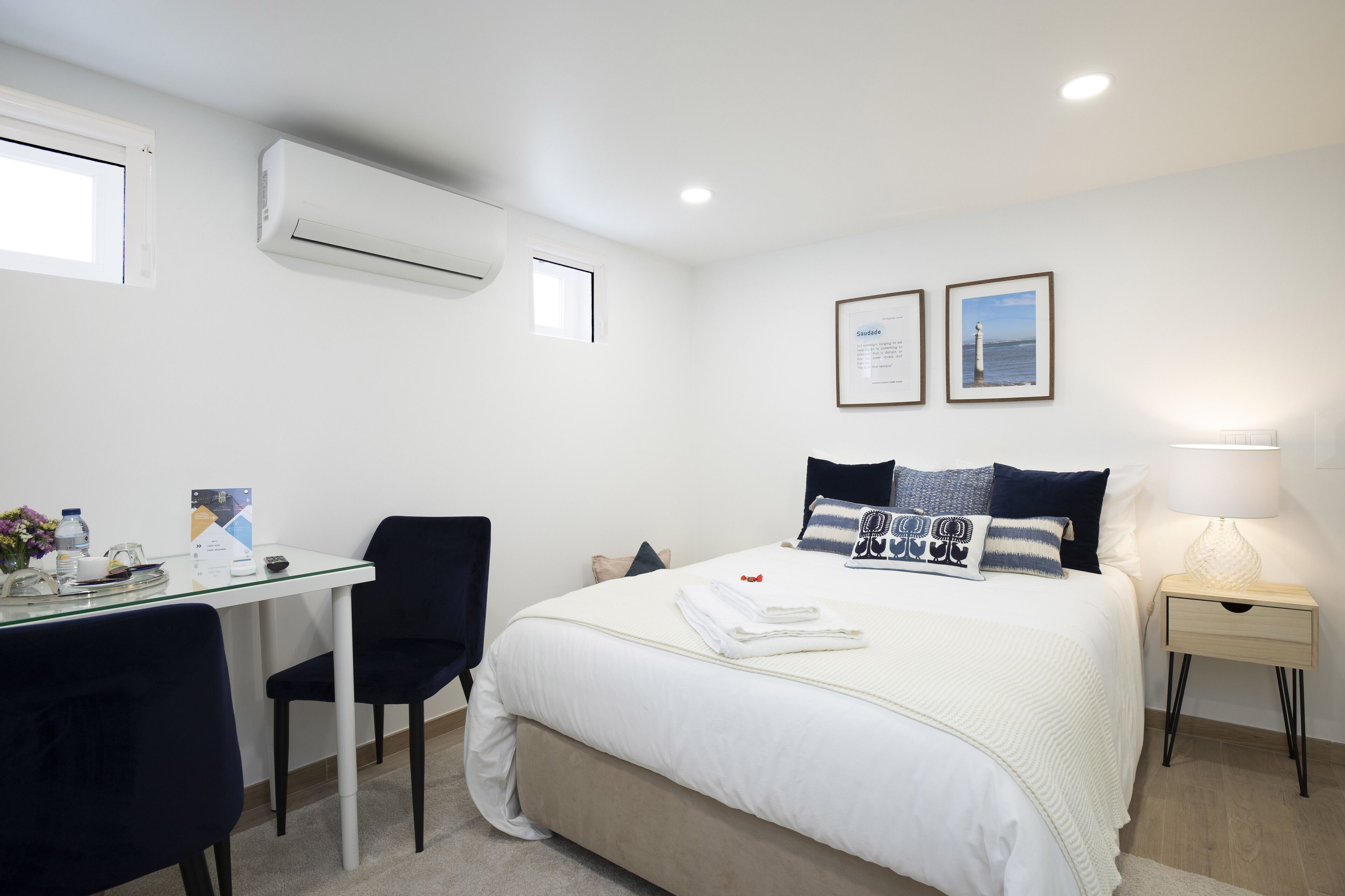 Habitación de hotel con cama, escritorio con silla, mesita de noche con lámpara y aire acondicionado.