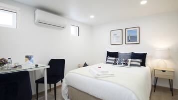 Habitación de hotel con cama, escritorio con silla, mesita de noche con lámpara y aire acondicionado.
