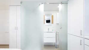A modern hotel room with a bed, a glass partition, a bathroom with a sink and mirror, and white cabinets. - Stupendo Airport Studio 1 (Lisboa)