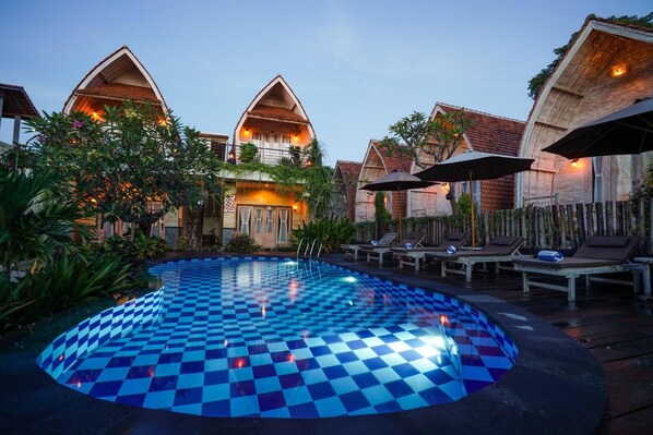 Une piscine à motifs à carreaux, entourée de chaises longues et de parasols, avec des bâtiments de style traditionnel en arrière-plan.