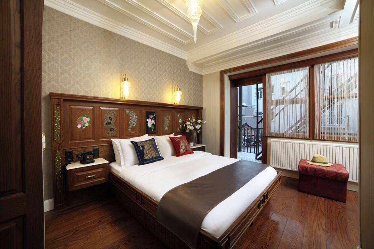 A hotel room with a large bed, wooden headboard, two bedside lamps, a nightstand, a window with a view, and a red ottoman.