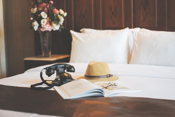 A bed with white sheets, a telephone, a book, a hat, and a vase of flowers.
