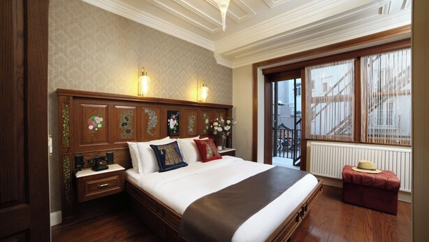 A hotel room with a large bed, wooden headboard, two bedside lamps, a nightstand, a window with a view, and a red ottoman.