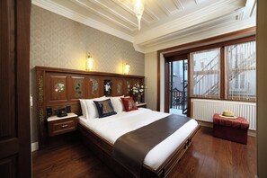 A hotel room with a large bed, wooden headboard, two bedside lamps, a nightstand, a window with a view, and a red ottoman.