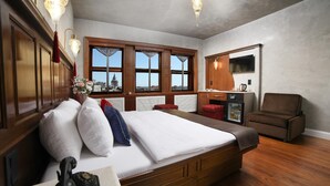 A hotel room with a large bed, wooden headboard, a small desk with a telephone, a chair, a television, and a view of a cityscape through large windows.