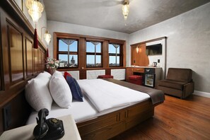 A hotel room with a large bed, wooden headboard, a small desk with a telephone, a chair, a television, and a view of a cityscape through large windows.