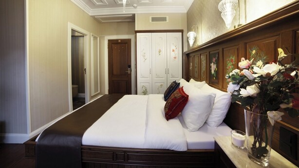 Quarto de hotel com uma cama grande, uma mesa de cabeceira com um vaso de flores, cabeceira de madeira e um guarda-roupa com decorações florais.