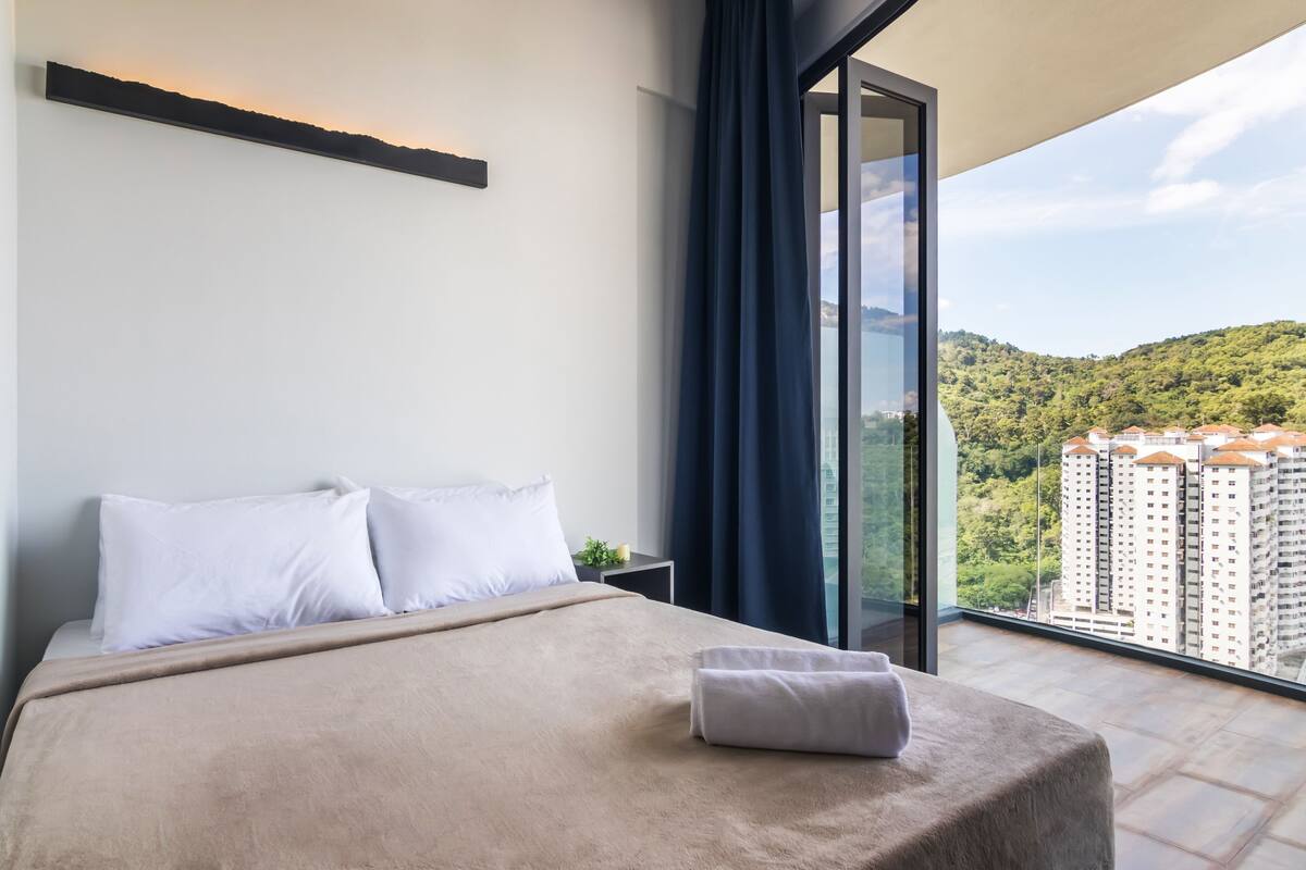 A modern hotel room with a large bed, a nightstand, and a view of a cityscape through floor-to-ceiling windows.