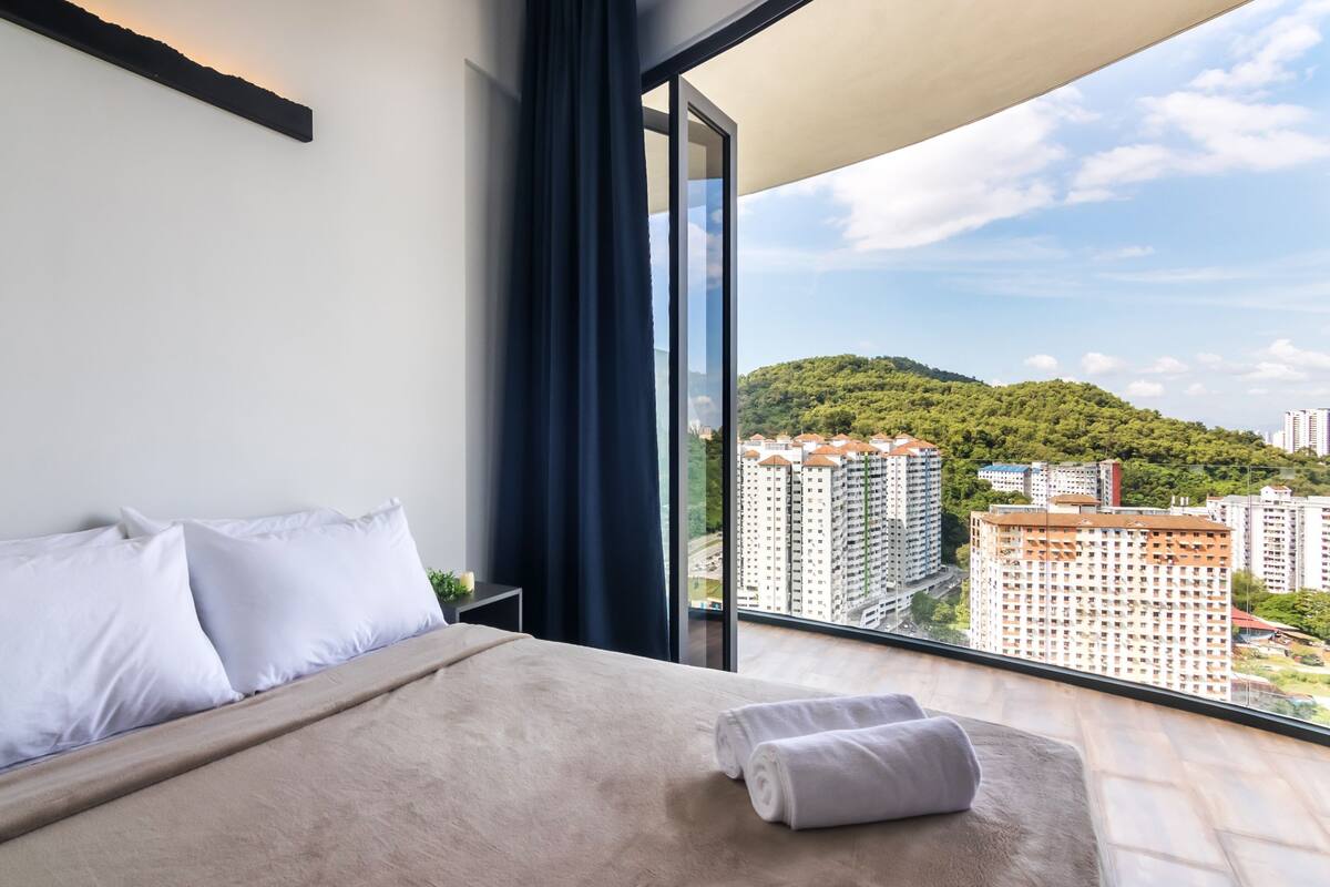 A hotel room with a large bed, a view of a cityscape, and a balcony with a view of a hill.