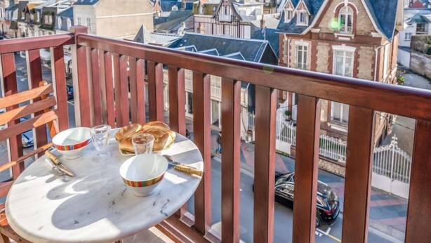 A balcony with a table set for two, overlooking a street with parked cars and traditional European-style buildings. - Apartment Le Hoc by Interhome (Deauville-Trouville)
