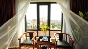 A room with a large window, two wooden chairs, a small table with a vase of flowers, and a potted plant. - Diamond Hotel (Ninh Binh)