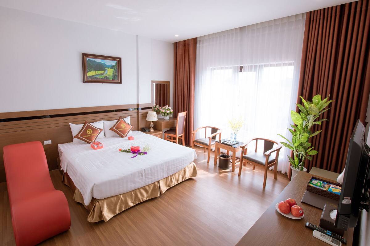 A hotel room with a bed, a desk with a computer, a chair, a small table, and a potted plant.