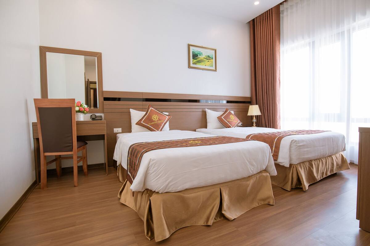 A hotel room with two beds, a desk, a chair, a mirror, and a window with curtains.