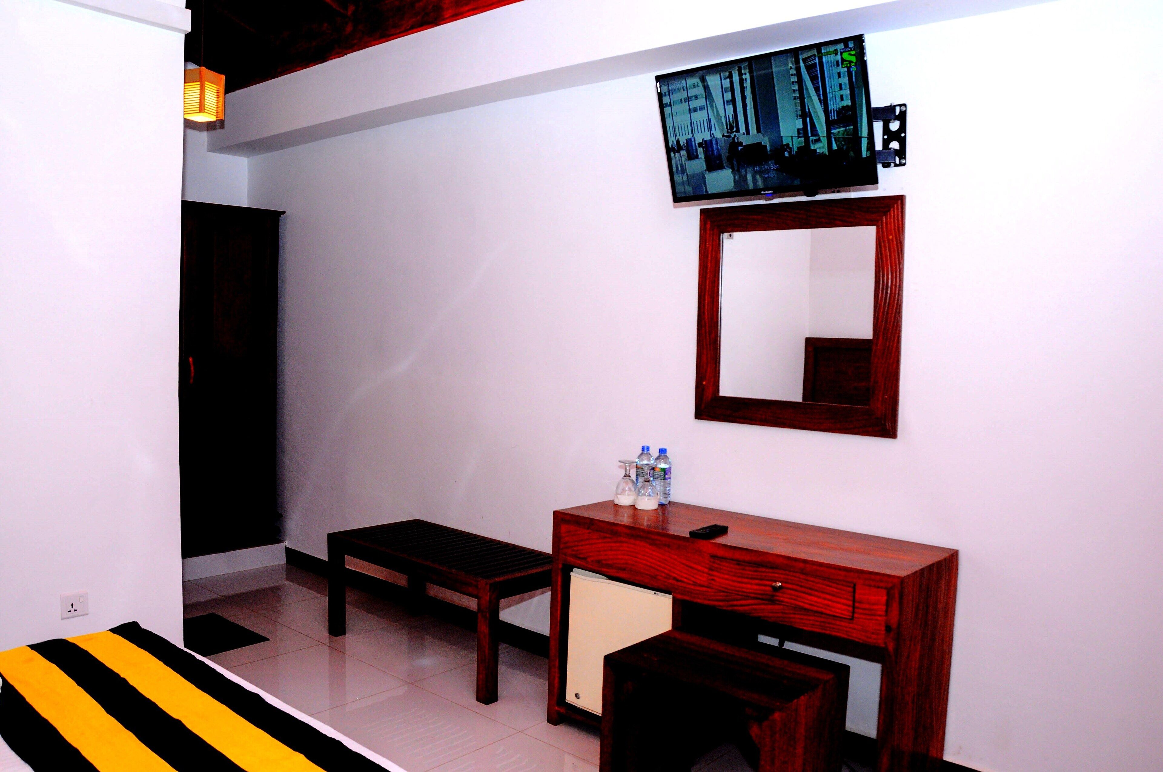 A hotel room with a wooden desk, a small refrigerator, a bench, and a television mounted on the wall.