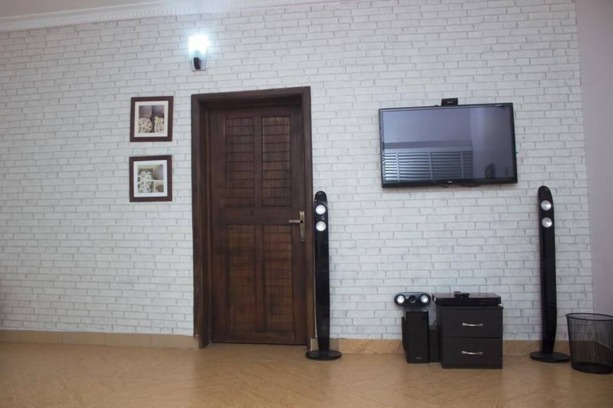 A room with a white brick wall, a wooden door, a flat-screen TV mounted above it, and a small table with drawers and speakers.