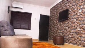 Ein Wohnzimmer mit einer Couch, einem Hocker und einem gemusterten Kissen. Eine Steinwand mit einem angebrachten Fernseher und einer Tür zu einem anderen Raum.