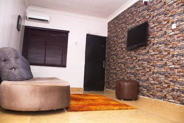 Un salón con un sofá, un reposapiés y un cojín estampado. Se observa una pared de piedra con un televisor montado y una puerta que da a otra habitación.
