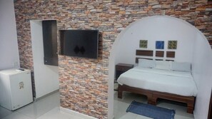 A bedroom with a stone wall, a bed with a white headboard, a TV mounted on the wall, and a small bedside table. - Studio 21 (Lagos)