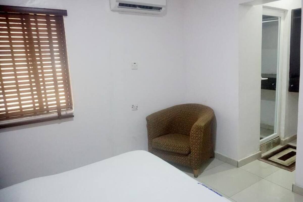 A small room with a single bed, a patterned armchair, a window with blinds, and a door leading to a closet.