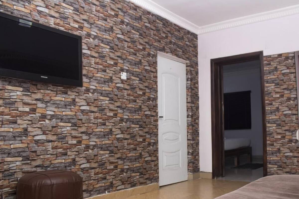 A room with a stone wall, a flat-screen TV mounted above a door, a brown ottoman, and a large bed with a brown rug.