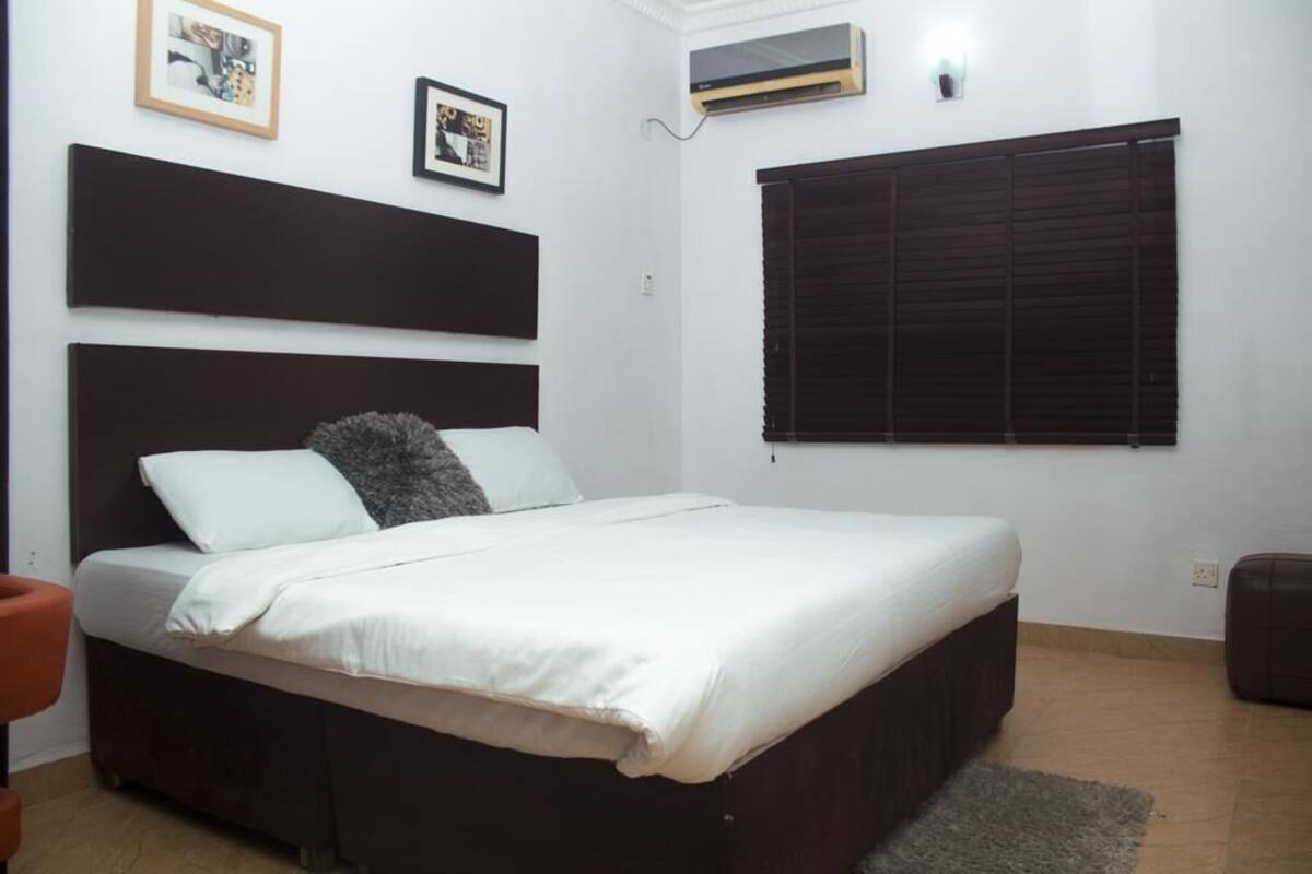 A bedroom with a bed, a headboard, a window with blinds, and a wall-mounted air conditioning unit.