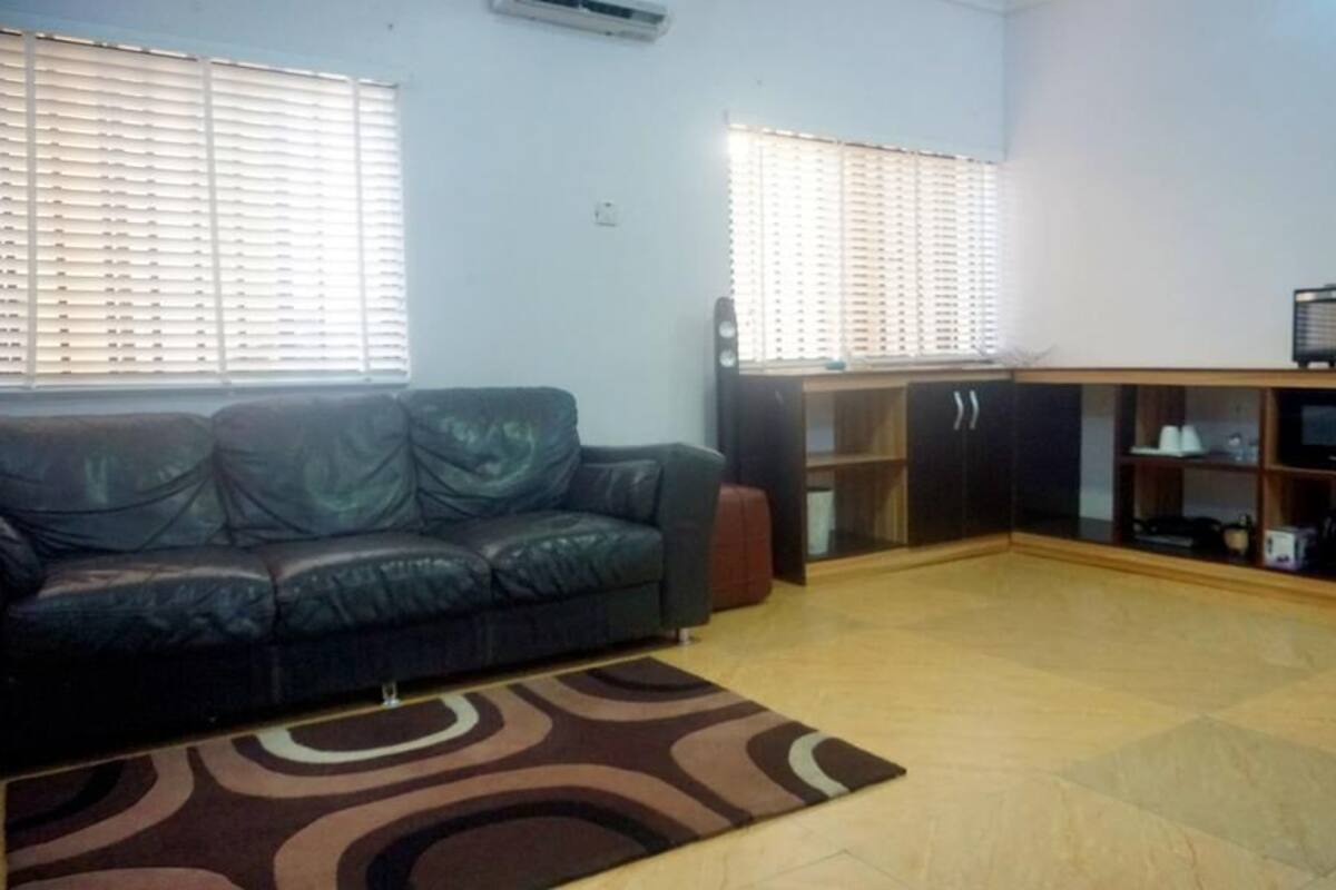 A living room with a dark leather sofa, a patterned rug, wooden flooring, a cabinet with shelves, and a microwave on a wooden cabinet.