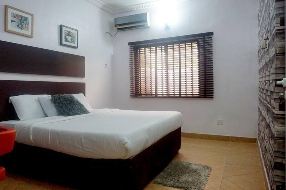 A bedroom with a bed, a red chair, a window with blinds, and a wall-mounted air conditioner.